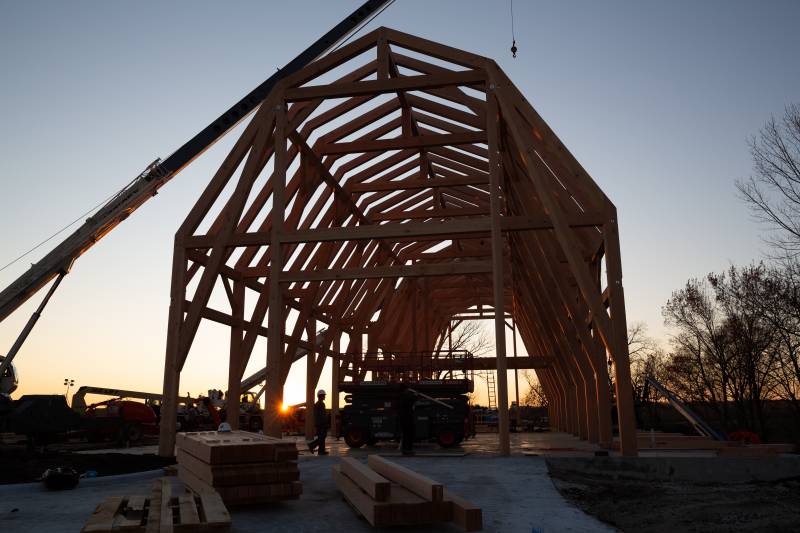 Timber framer walks by the setting Iowa sun with the frame behind him